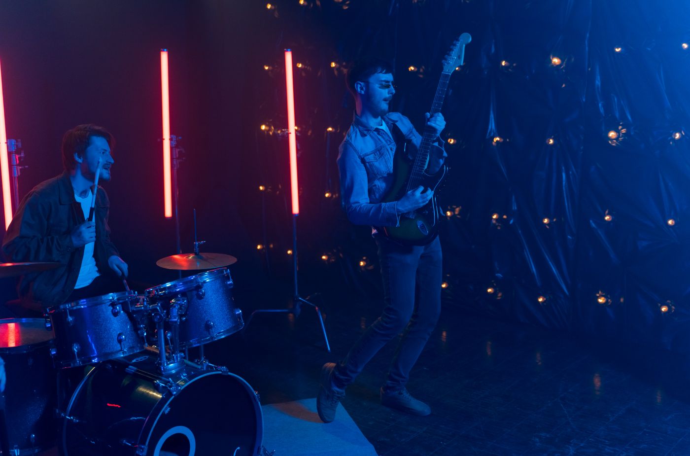 A guitarist and drummer in a rehearsal room with colored light